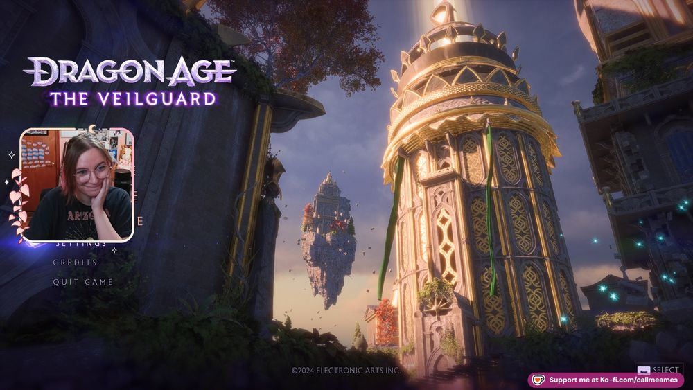 Ames' facecam over the main screen for Dragon Age Inquisition. They are smiling with a hand pressed to their cheek, looking very excited! 