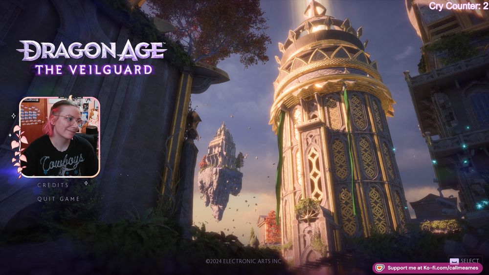 Ames' facecam over the main menu for Dragon Age: The Veilguard. Ames has her head tilted and is smiling. Their hair is pulled back and they are wearing a black shirt that says cowboys on it it white font. In the upper right corner of the screen is a cry counter showing that ames has cried two times at veilguard already.