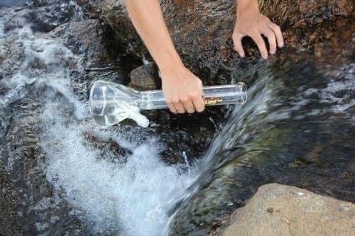 a person holding a clear and large bong infront of a stream of water in nature.