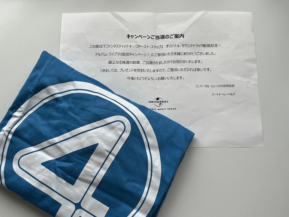 「キャンペーンご当選のご案内
この度は「『ファンタスティック4：ファースト・ステップ』オリジナル・サウンドトラック配記念！アルバム・ライブラリ追加キャンペーン！」にご参加いただき誠にありがとうございました。
厳正なる抽選の結果、ご当選されましたのでお知らせいたします。
つきましては、プレゼントを同封いたしますので、ご査収いただければ幸いです。
今後ともどうぞよろしくお願いいたします。
ユニバーサルミュージック合同会社パートナーレーベルズ」と書かれた紙と、4のロゴの入ったTシャツが折り畳まれて写ってる。