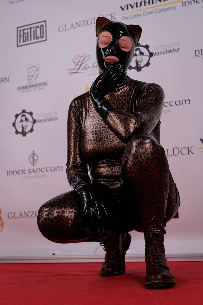 ToyDolly in a shiny leopard print zentai with black latex gloves and kitty hood. They are in a crouched position and considering the camera as prey