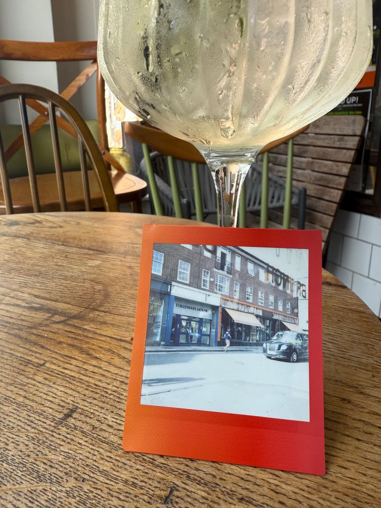 A Instax image depicting a building with the text “threeways house” leaning against a frosty Hugo spritz on a wooden table. 