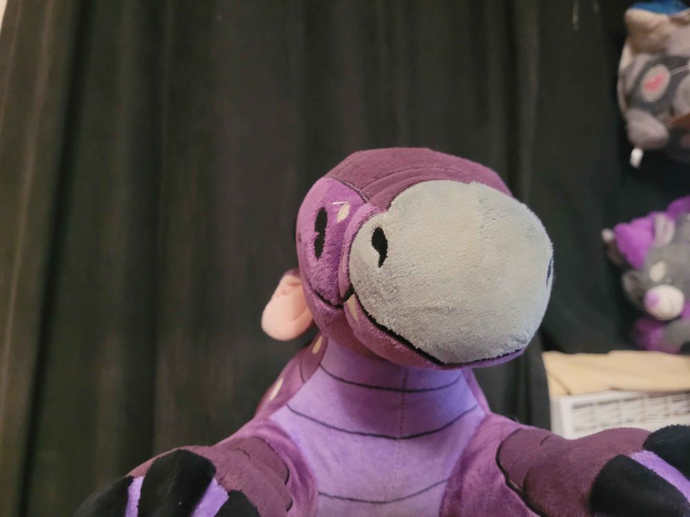 Photo of a purple dragon plushie looking at the camera, the dragon has black claws, a grey nose, and white spots on his face and neck.