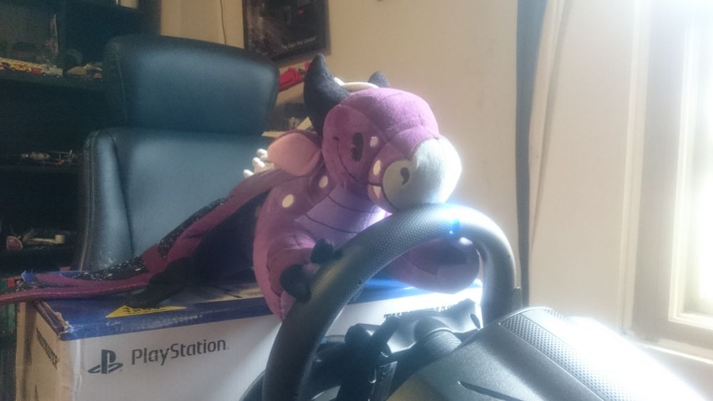 Photo of the same dragon plushie as the previous images, this time he is sitting on a box with his front claws holding onto a gaming steering wheel. (His ass does NOT have his drivers license!)