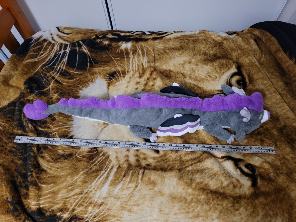 A photo of the same dragon plush as the previous images, this time he is layed out across a blanket with a meterstick next to him showing him to be 1 meter long.