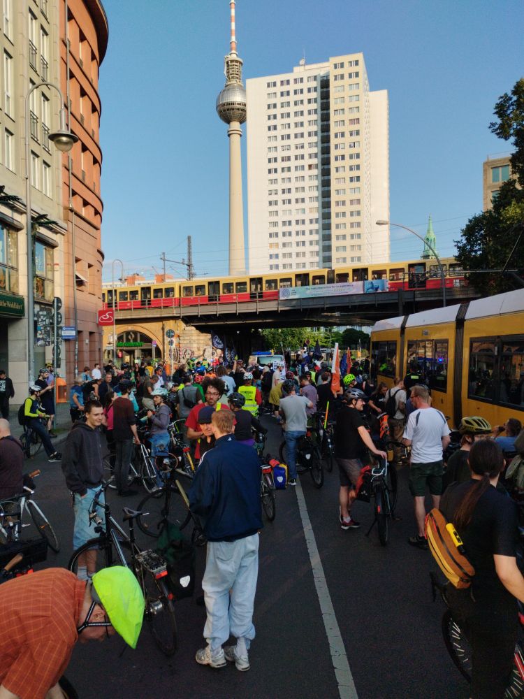 Fahrradkundgebung mit vielen bunten Teilnehmenden. Im Hintergrund eine S-Bahnbrücke mit Bahn, der Berliner Fernsehturm und ein Hochhaus.
