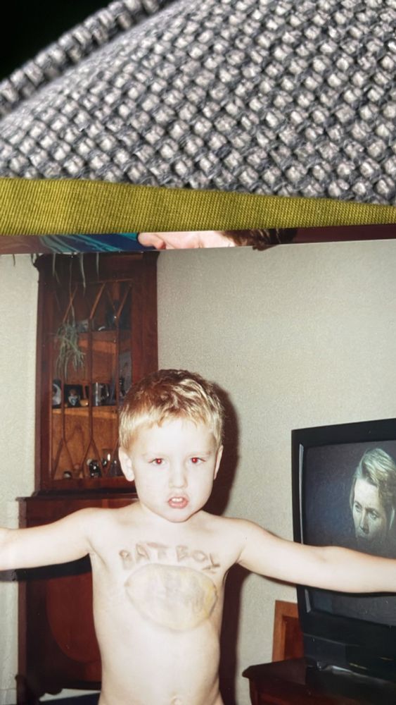 A picture of me as a boy, maybe 7 or 8, topless and arms outstretched with the Batman logo and the words Bat Boy painted on my chest as I stare down the camera. Behind me on a CRT TV from the 90s is the head of an woman unknown blonde woman, maybe in her 40s