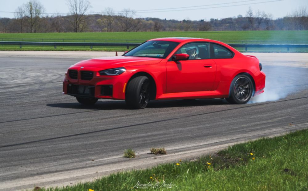 A red BMW M2 at yaw with smoke coming from the rear tires. The driver is giving a peace sign to the photographer.