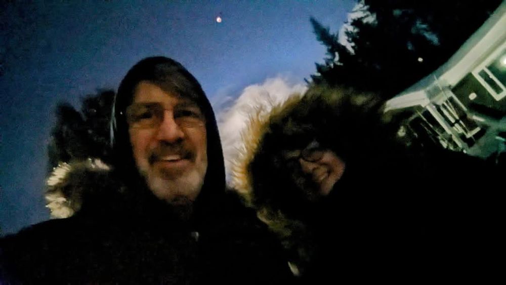 man and woman in parkas smiling with the eclipse behind them