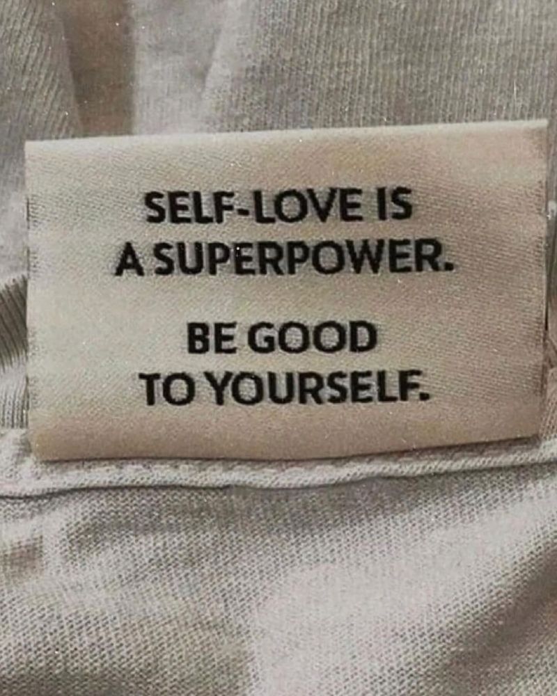 A close-up photograph of a small fabric or cloth label with black text printed on a light beige background. The text reads "SELF-LOVE IS A SUPERPOWER. BE GOOD TO YOURSELF." in capital letters across two lines. The label appears to be photographed against a textured light gray fabric background.