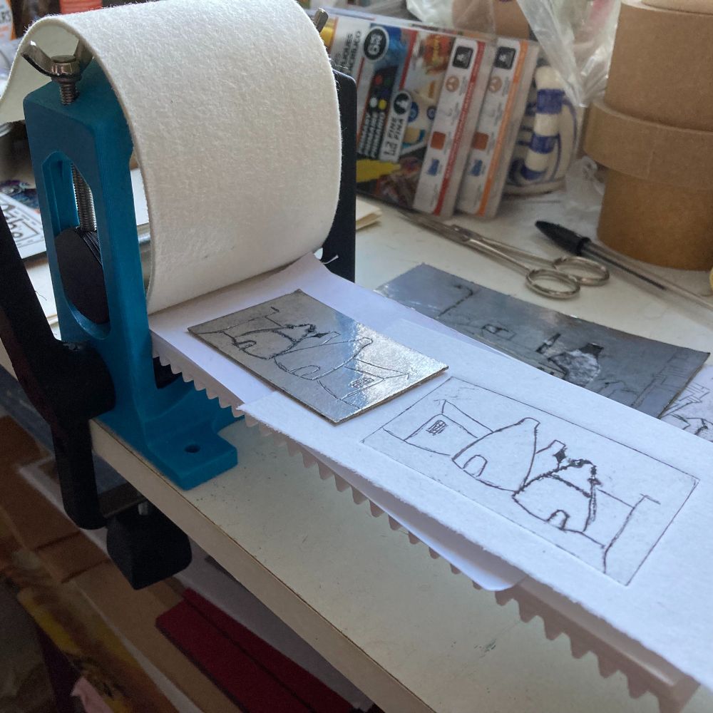 This one shows the tiny roller press, it has a light blue plastic 3-d printed frame, and is clamped to my desk. The long white press bed has a serrated underside, the teeth run through the mechanism when the handle is turned. On the press bed is the print of the made up group of three bottle kilns and its foil plate, and a white felt blanket is folded back over the top of the press. There are scissors, pens, a plie of various rolls of tape and packets at the back of the image.