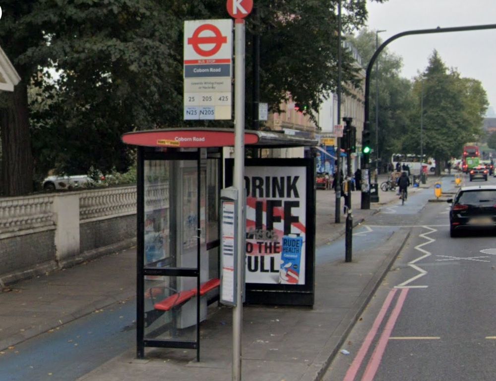 A view from the road of a bus stop, on an area of pavement between the road a bike lane, painted blue, and the normal pavement the other side of that. It has a bus shelter, the side near us and most of the back glazed, but the side of bus shelter away from us is a solid panel with a big advert over all of it. Behind this is an area of pavement where idiots like to step into the bike lane without looking, and you can predict this because they're hidden behind the big advert. 