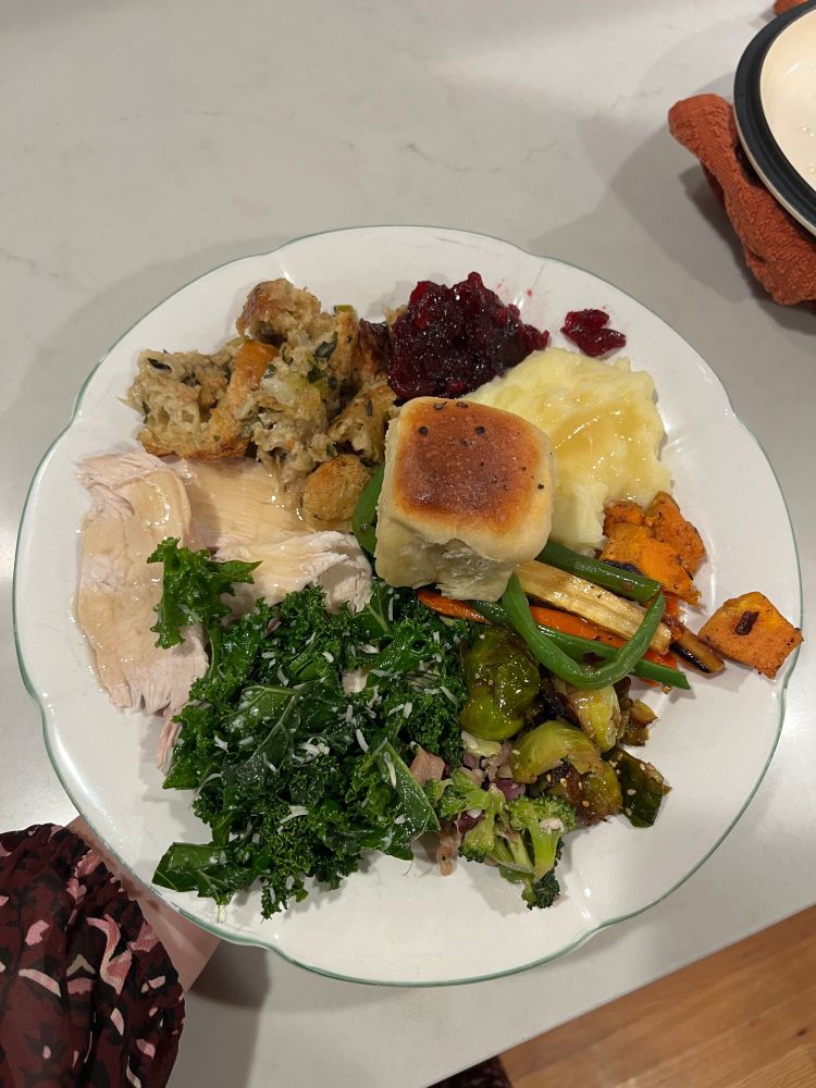 A stunning Thanksgiving plate with stuffing, mashed potatoes, cranberry sauce,  roll, turkey and gravy, kale salad, green beans, carrots and parsnips, sweet potatoes, Brussels sprouts, and broccoli salad 