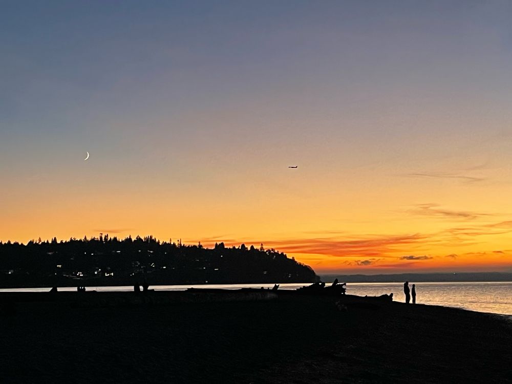 Moon and airplane in the sky with the sunset glowing orange over the beach