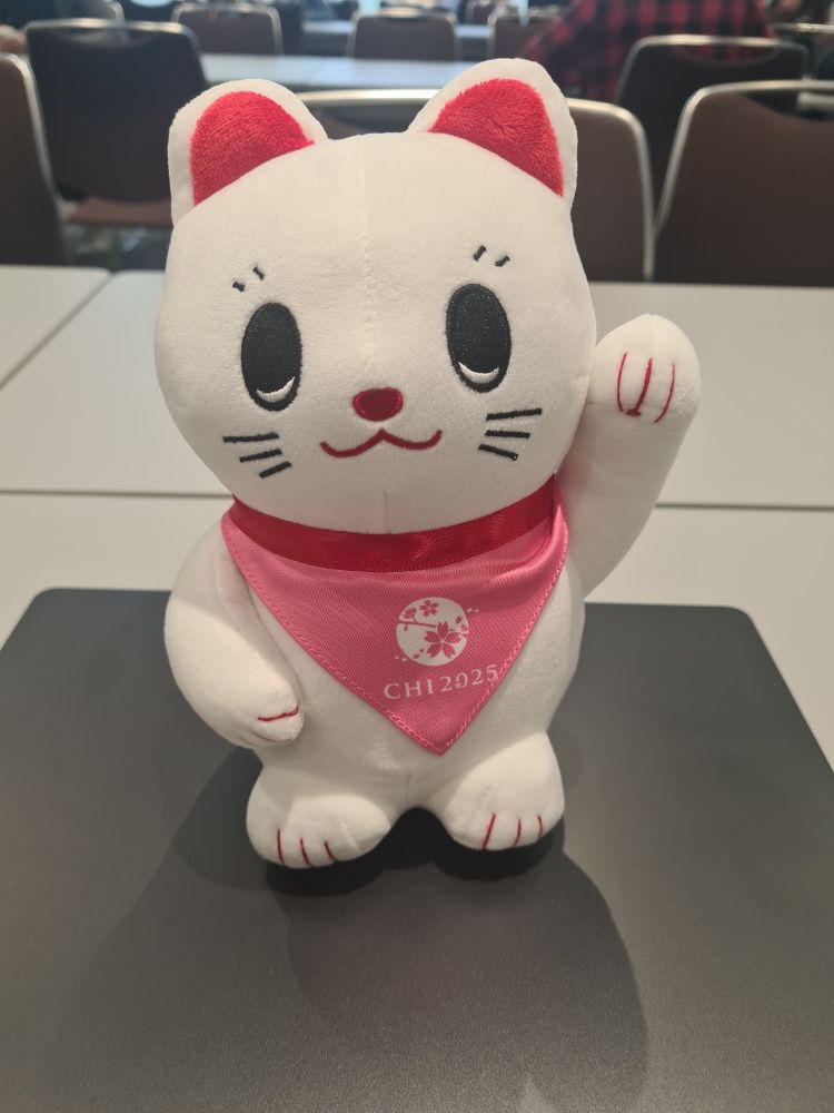 A white maneki-neko plushie with a CHI2025 scarf looking extra cute