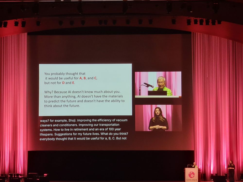 View of an auditorium stage with slides showing in the centre and Wakamiya-san and a sign-language interpreter showing on the sides.