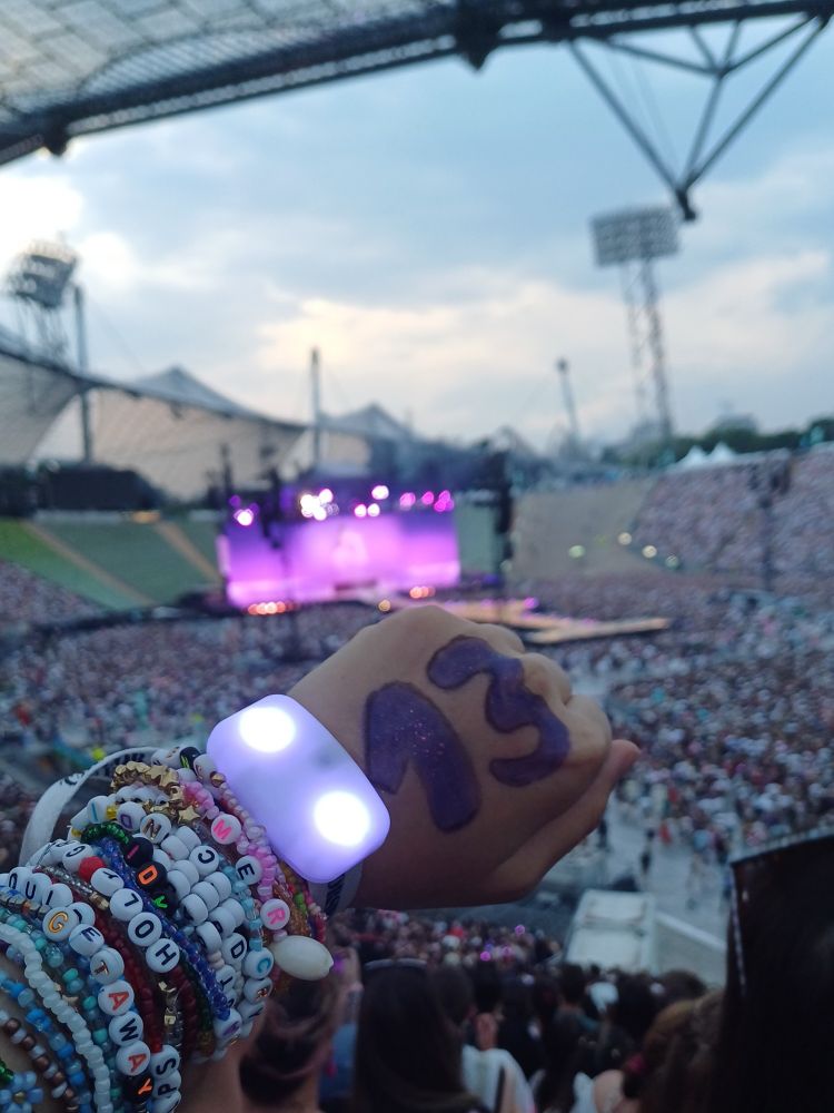 me stretching out an arm at the eras tour with a 13 painted on my hand and lots of friendship bracelets on my hand while taylor is performing enchanted