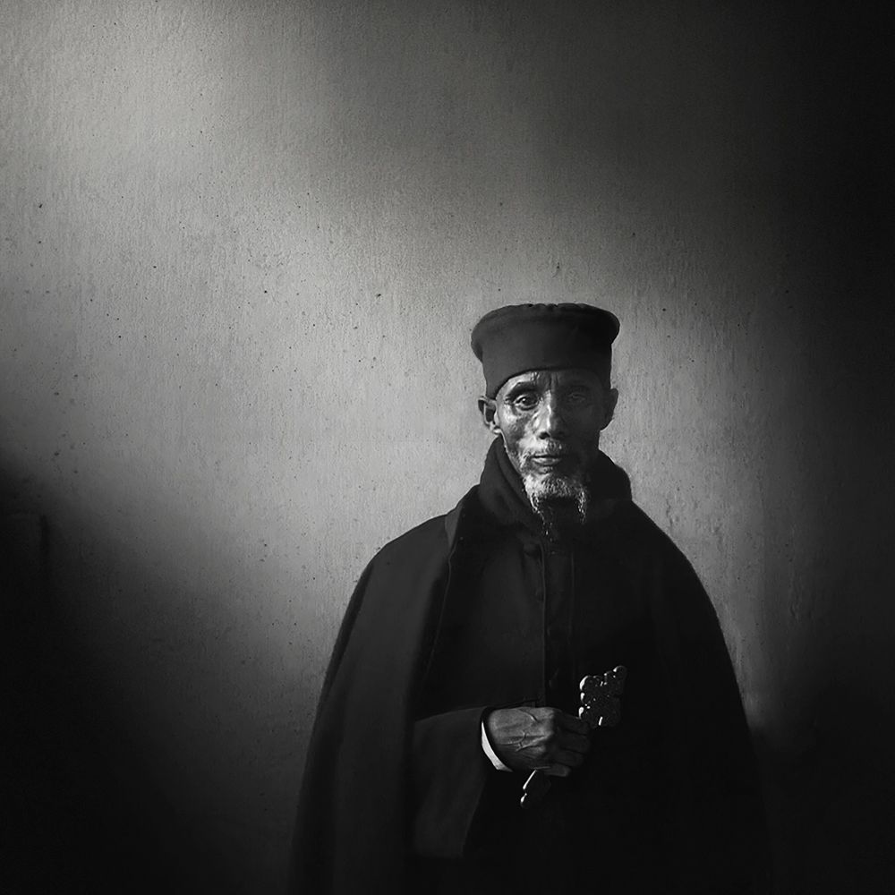 An elderly Ethiopian Priest stands against a bare wall, draped in dark robes and holding a cross. The light falls softly across his face, revealing depth, history, and quiet dignity in the stillness of his presence