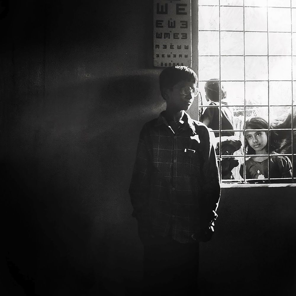 A boy stands beneath an eye chart, cast in soft shadow. Light pours through a barred window beside him where a young girl looks in, her expression quiet and curious. The scene feels like a moment between worlds.