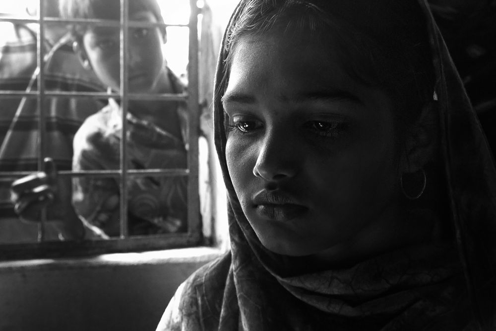 A young girl in Bangladesh sits in deep shadow, her face softly lit with quiet emotion. Behind her, a boy peers through metal bars, his eyes searching. The light and framing evoke a feeling of separation, longing, and quiet resilience