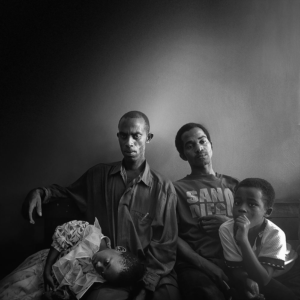 Three figures sit together — two men, one with a child asleep on his lap, the other resting back with a calm gaze. A fourth child leans forward, deep in thought. The image is rich with unspoken bonds, fatigue, and waiting.