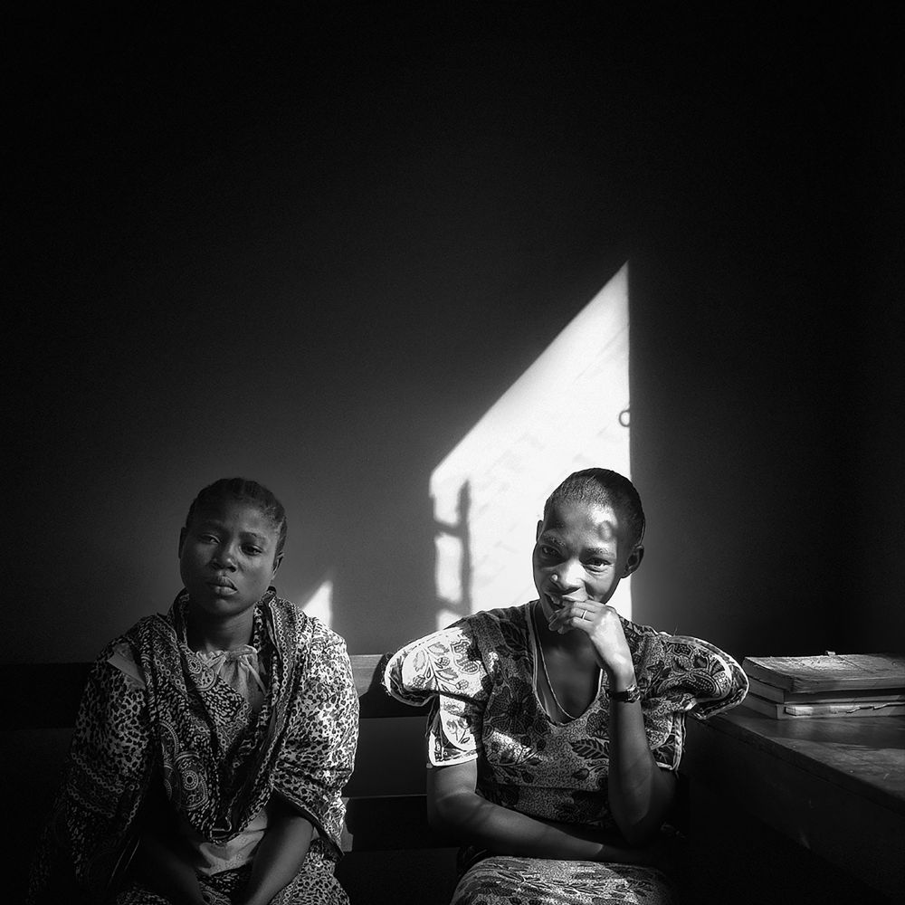 Two women sit side by side, one gazing away in thought, the other with a faint smile, hand resting under her chin. A triangle of bright light on the wall casts long shadows, giving the scene both tension and intimacy.
