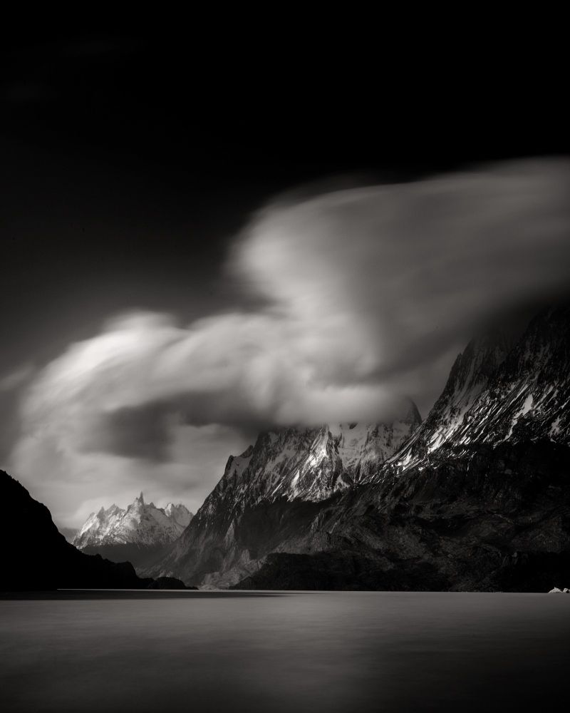 Dramatic black and white long exposure photograph of Lago Grey in Patagonia. Towering, snow-covered mountains rise sharply in the background, partially obscured by streaking, fast-moving clouds. The lake in the foreground appears smooth and glassy, adding a sense of stillness to the dynamic sky above. The moody lighting enhances the stark contrast between light and shadow, emphasizing the rugged texture of the landscape