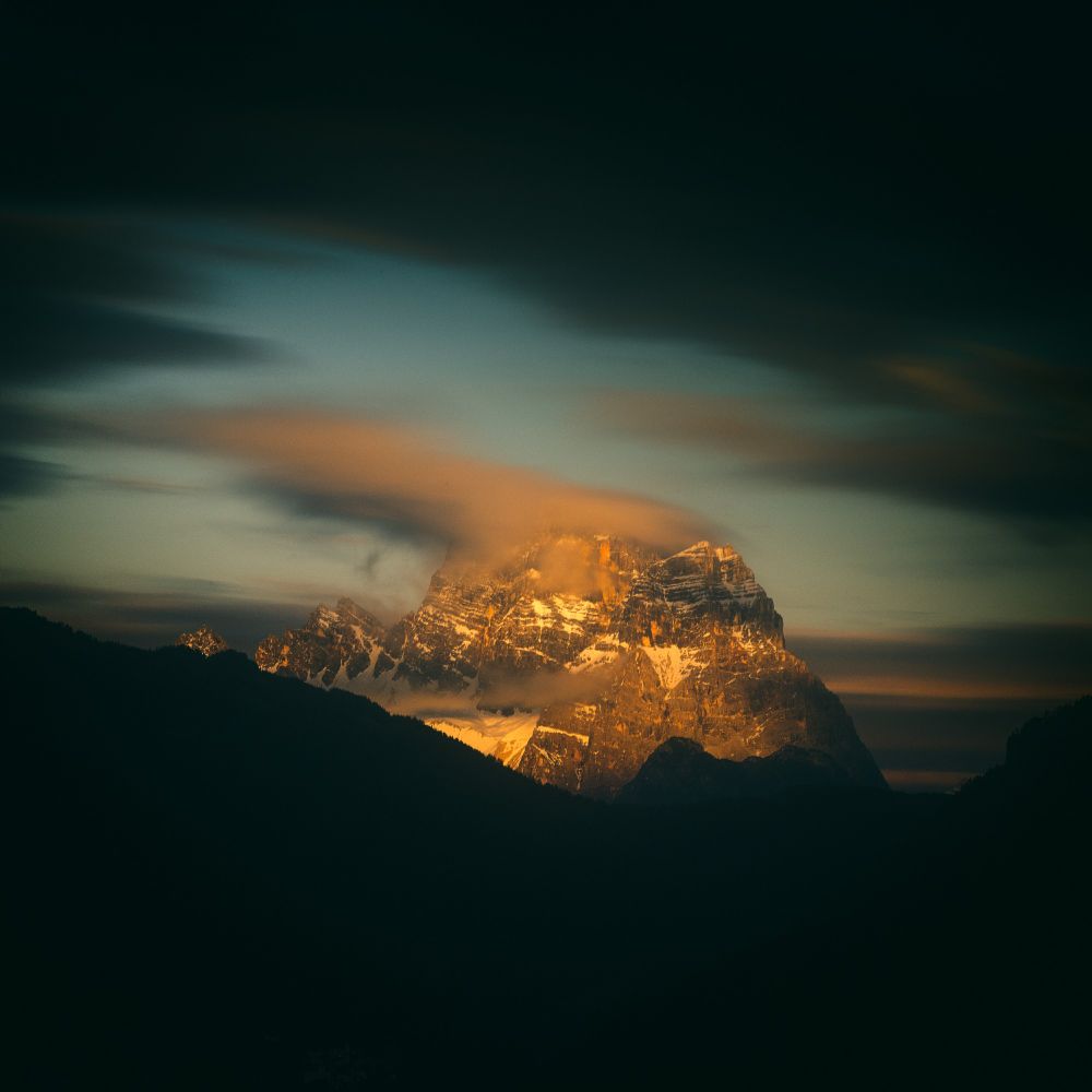 This image captures a stunning mountain peak in the Dolomites, Italy, during what appears to be either sunrise or sunset. The golden light casts a dramatic glow on the rugged rock face, highlighting the sharp, angular ridges and the remnants of snow clinging to the slopes. Wispy clouds partially obscure the peak, adding a sense of mystery and movement to the scene.