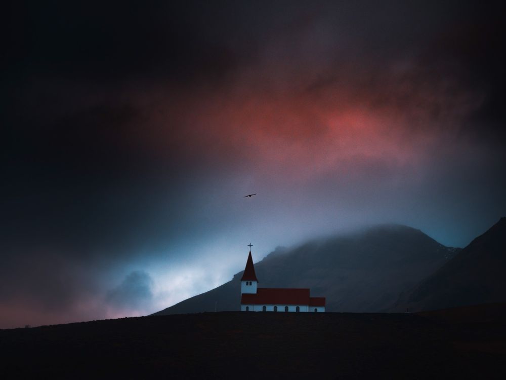 A solitary white church with a red roof and steeple stands on a dark hillside beneath dramatic, moody skies in Vík, Iceland. A single bird soars above as mist and low clouds partially obscure the surrounding mountains, with a hint of red and blue light breaking through the stormy atmosphere