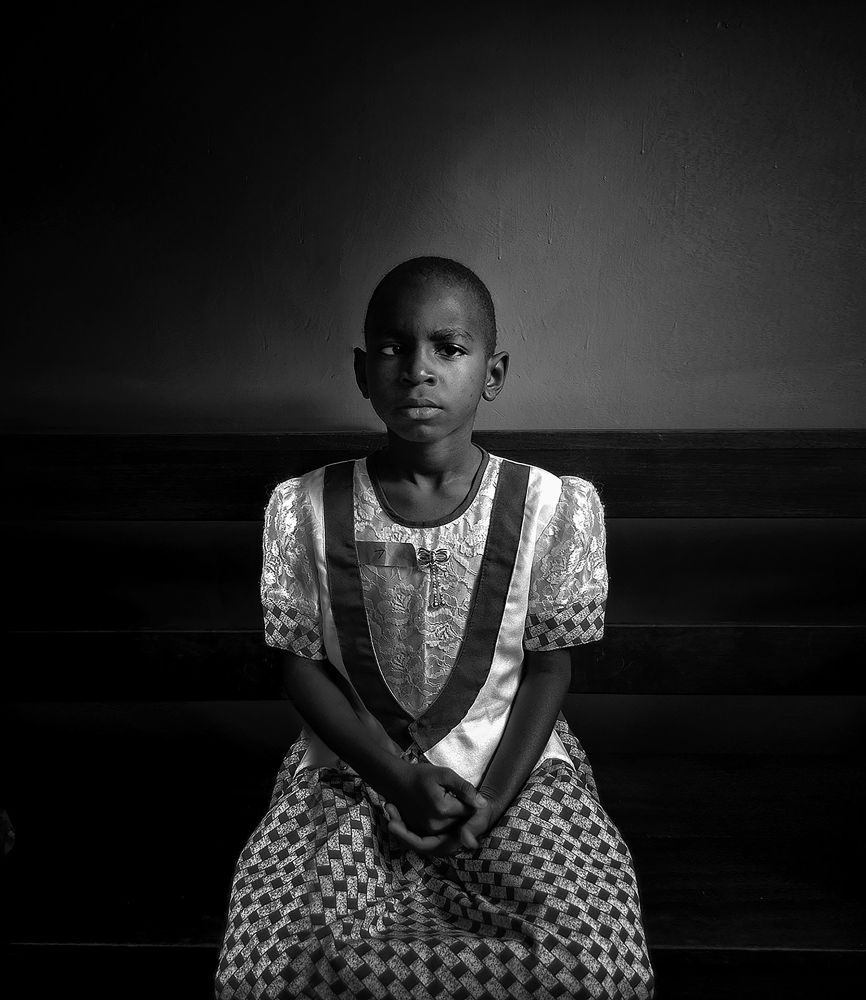 A Tanzanian girl in a patterned dress sits upright on a wooden bench, hands gently clasped. The room is stark, the light focused, isolating her in a moment of stillness. Her gaze is calm but carries the weight of silence.