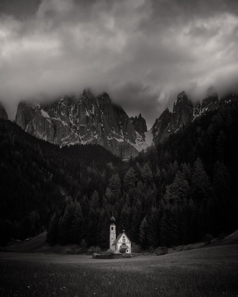 A small, picturesque church—St. Johann in Ranui—sits in a peaceful meadow at the edge of a dense pine forest in Val di Funes, Italy. Towering, jagged peaks of the Dolomites rise dramatically behind it, partially shrouded in mist and low-hanging clouds. The black-and-white composition enhances the moody, ethereal atmosphere of the alpine scene.