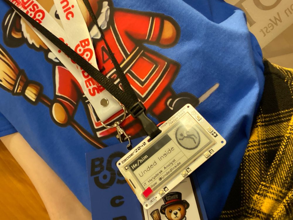 Selfie of my torso. I’m wearing a blue bsides T-shirt - with a picture of a teddy bear wearing a beefeater uniform - and an epaper badge on a lanyard around my neck with my handle, logo, and pronouns on