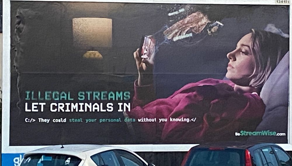 Anti piracy billboard showing a woman laying down looking at her phone, with an ominous hand above her. The billboard reads “Illegal streams let criminals in” and the subtitle says “C:/> They could steal your personal data without you knowing.</“