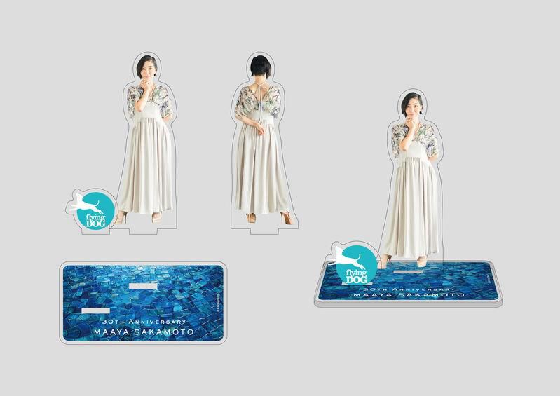 An acrylic standee of Maaya Sakamoto in a floral top and long white skirt posing with her hand under her chin. Instead of the same image mirrored on both sides, one side shows her front and the other shows her back. The base of the stand is a sapphire blue and also includes the Flying Dog logo