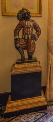 A statue of a black African. The statue is on a black pedestal with gold trim. The statue is kneeling on one knee and wearing a red and gold suit and a turban.