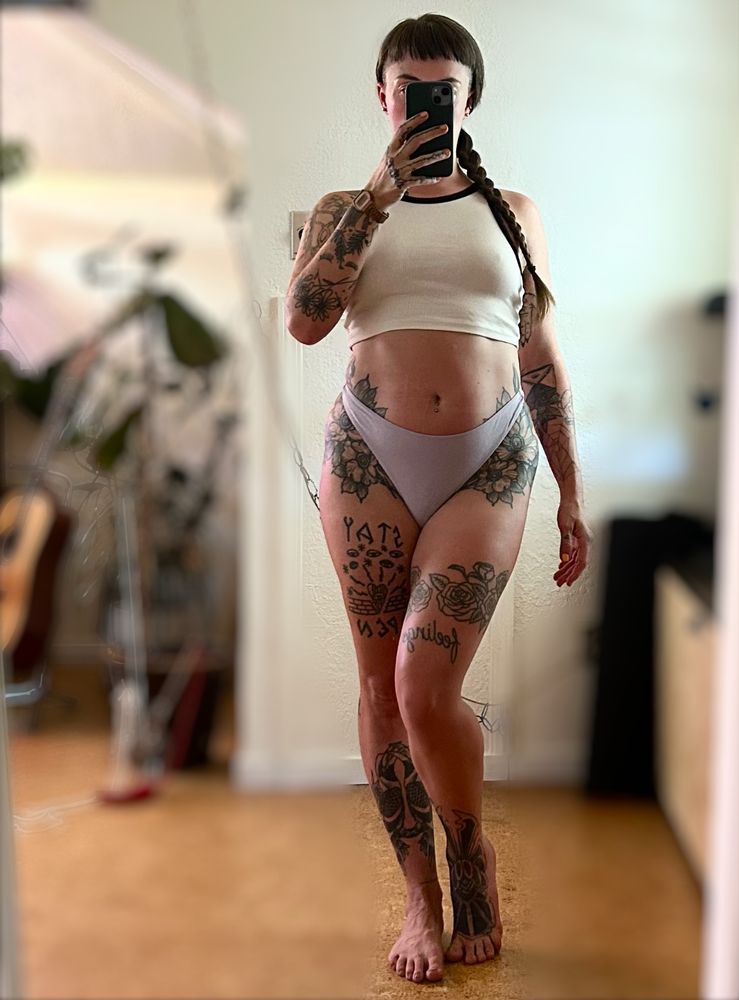 Rae stands in front of a mirror in a white cropped tank top and a lilac thong. Her hair falls to the side in a braid. 