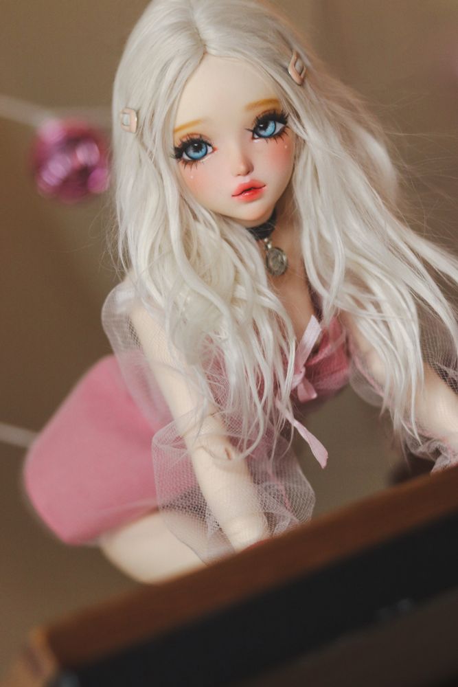 A ball-jointed doll with cream skin, white-blonde hair and blue eyes posed crawling toward the edge of the table. She’s wearing a pink dress with translucent sleeves. 

