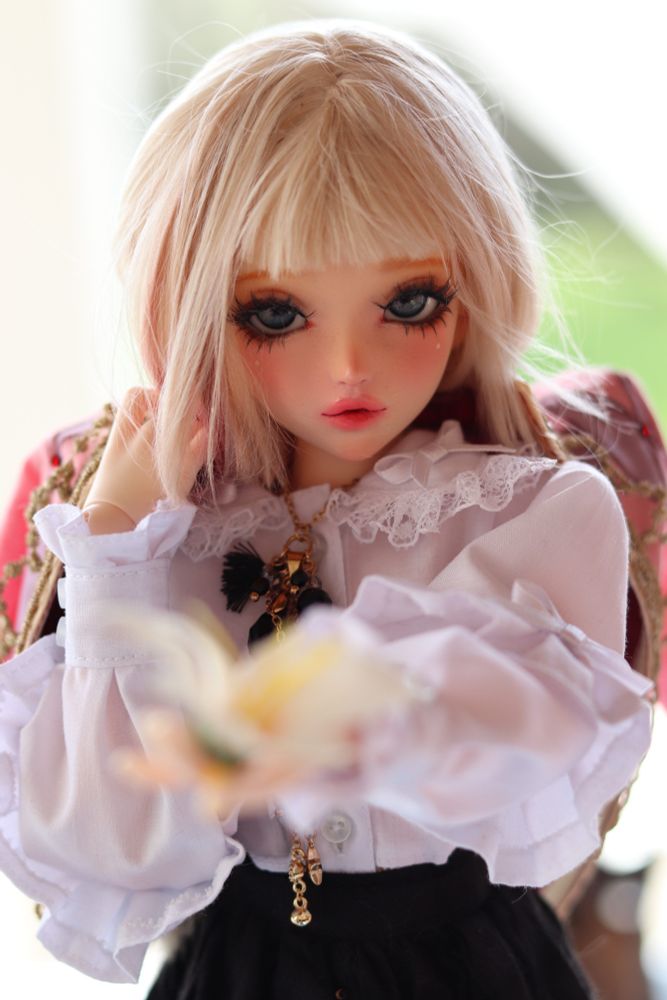 A ball-jointed doll with cream skin, blonde hair, and blue eyes posed extending one hand with a flower in it. 