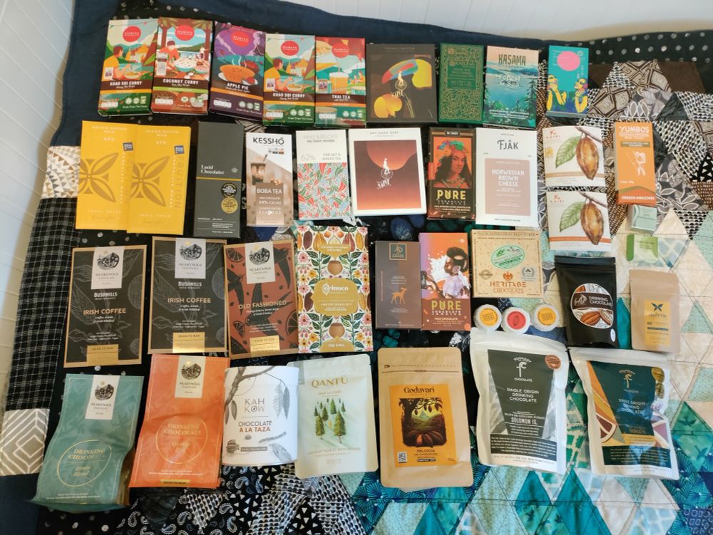 A few dozen bars of craft chocolate and bags of drinking chocolate from various craft chocolate companies all spread out nearly on a blanket