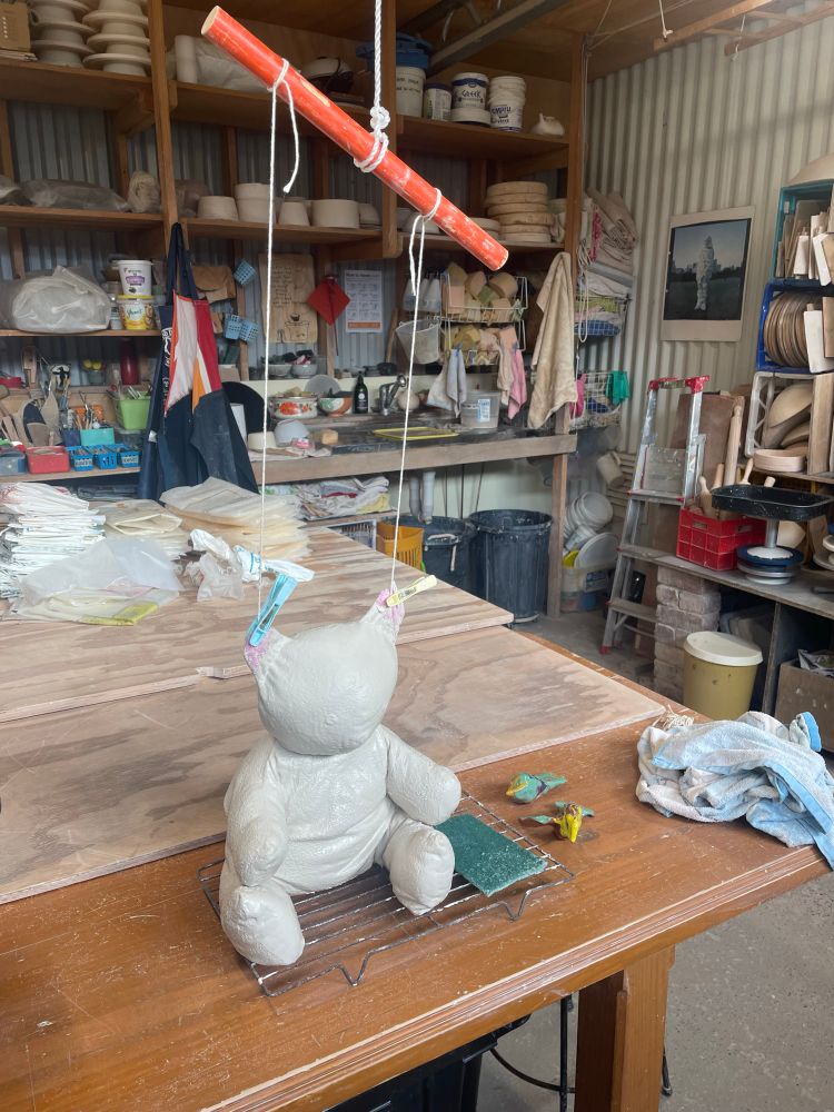 a teddy bear sitting on a table. it has been dipped in white liquid clay and has strings attached to its ears holding it up