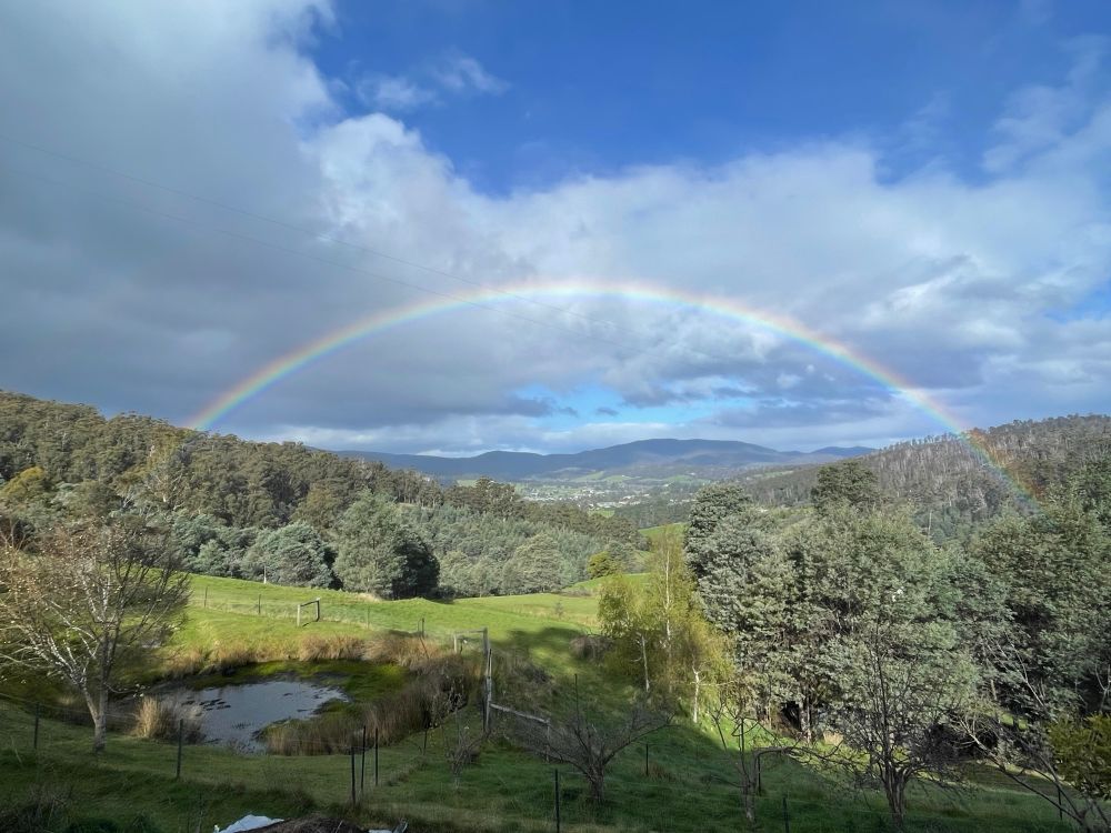 looking out into a green valley full of trees, the sky is blue with fluffy white clouds and a rainbow stretches over the valley 