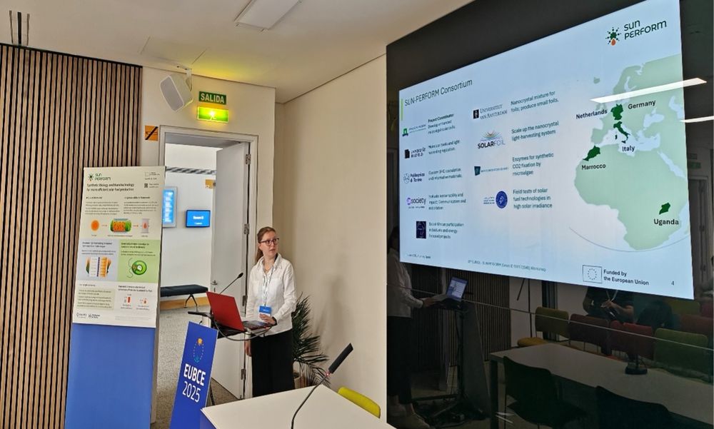 A person presenting a slide displaying the logos and locations of consortium members involved in a project on alternative biofuels.