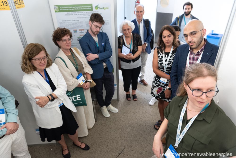 A person at a conference, surrounded by a small group of people, presents a poster about a project developing alternative fuels for aviation. 