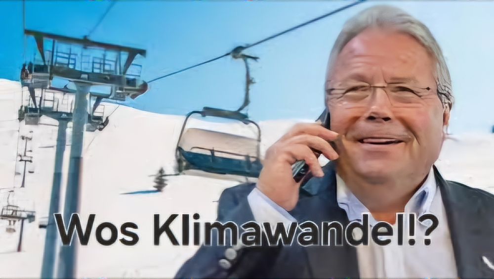 Eine Person mittleren Alters (Franz Hörl) mit grauen Haaren und Brille spricht draußen im Schnee mit einem Handy. Im Hintergrund sind ein Skilift und verschneite Berge zu sehen. Unten im Bild steht in großer Schrift der sarkastische Text: „Wos Klimawandel!?“.
