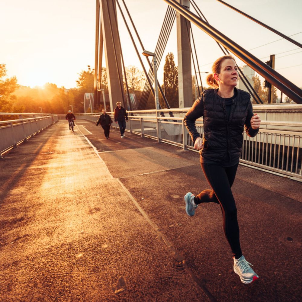 Beate Meindl-Reisinger joggt über eine Brücke bei Sonnenuntergang. Sie trägt eine schwarze Sportjacke, schwarze Leggings und blaue Laufschuhe. Ihre Haare sind zu einem Zopf gebunden, und sie wirkt konzentriert. Im Hintergrund gehen zwei Personen und ein Radfahrer über die Brücke. Die tiefstehende Sonne wirft ein warmes, goldenes Licht auf die Szene.