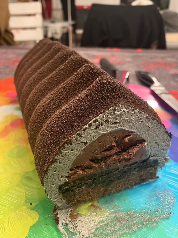 Gâteau en forme de bûche. De l'extérieur vers l'intérieur : un flocage marron, une mousse de sésame noir qui a donc une couleur grise, un crémeux au chocolat en demi-lune posé sur une couche croustillante au chocolat et sésame noir et un biscuit au chocolat.