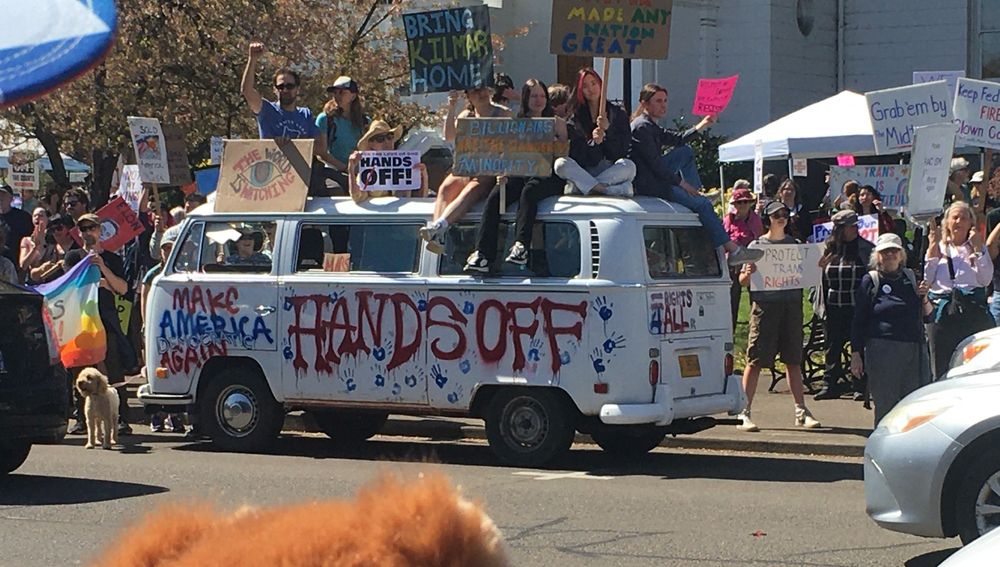 A van parked at the #HandsOff2025 rally in Corvallis, OR, painted with slogans - eg 'Make America Democratic Again'. A large number of protestors with signs surround the van, including on its roof.