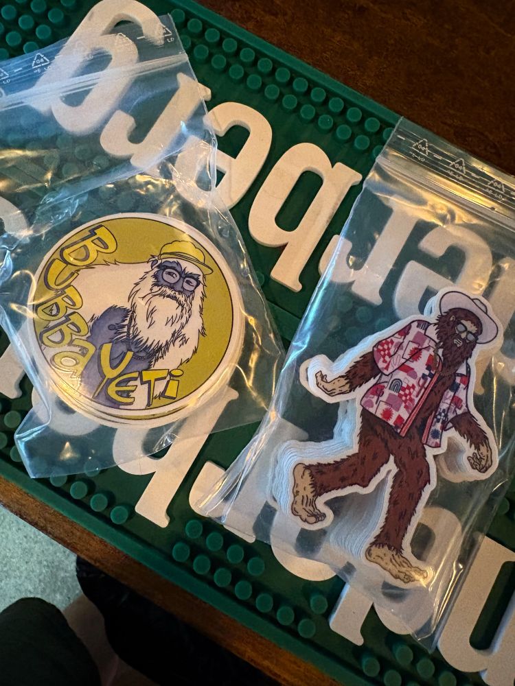 A pair of stickers. One is circular, yellow and gold, with a picture of Big Foot wearing a hat and glasses. The second is Big Foot walking wearing a floral shirt, glasses and a cowboy hat. 