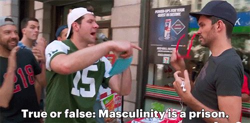 A gif of Billy Eichner, a white man, wearing a football jersey and a backwards white ball cap surrounded by a group of similarly dressed men. He is holding a microphone and asking another man wearing a black t shirt and black ball cap “true or false: masculinity is a prison.”