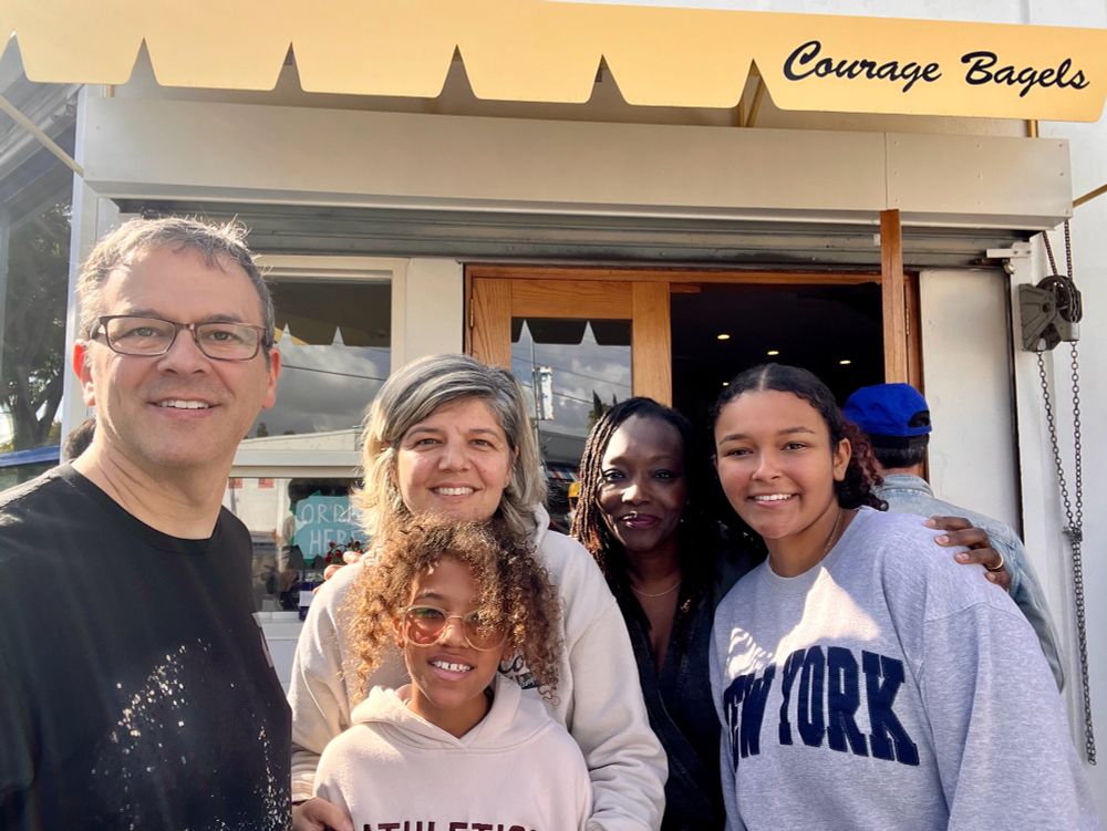 A few kids and adults posing together in front of a restaurant whose canopy reads “Courage Bagels”